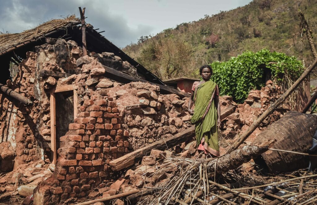 Woman stands in front of a destroyed house after a disaster in rural India, depicting struggle.