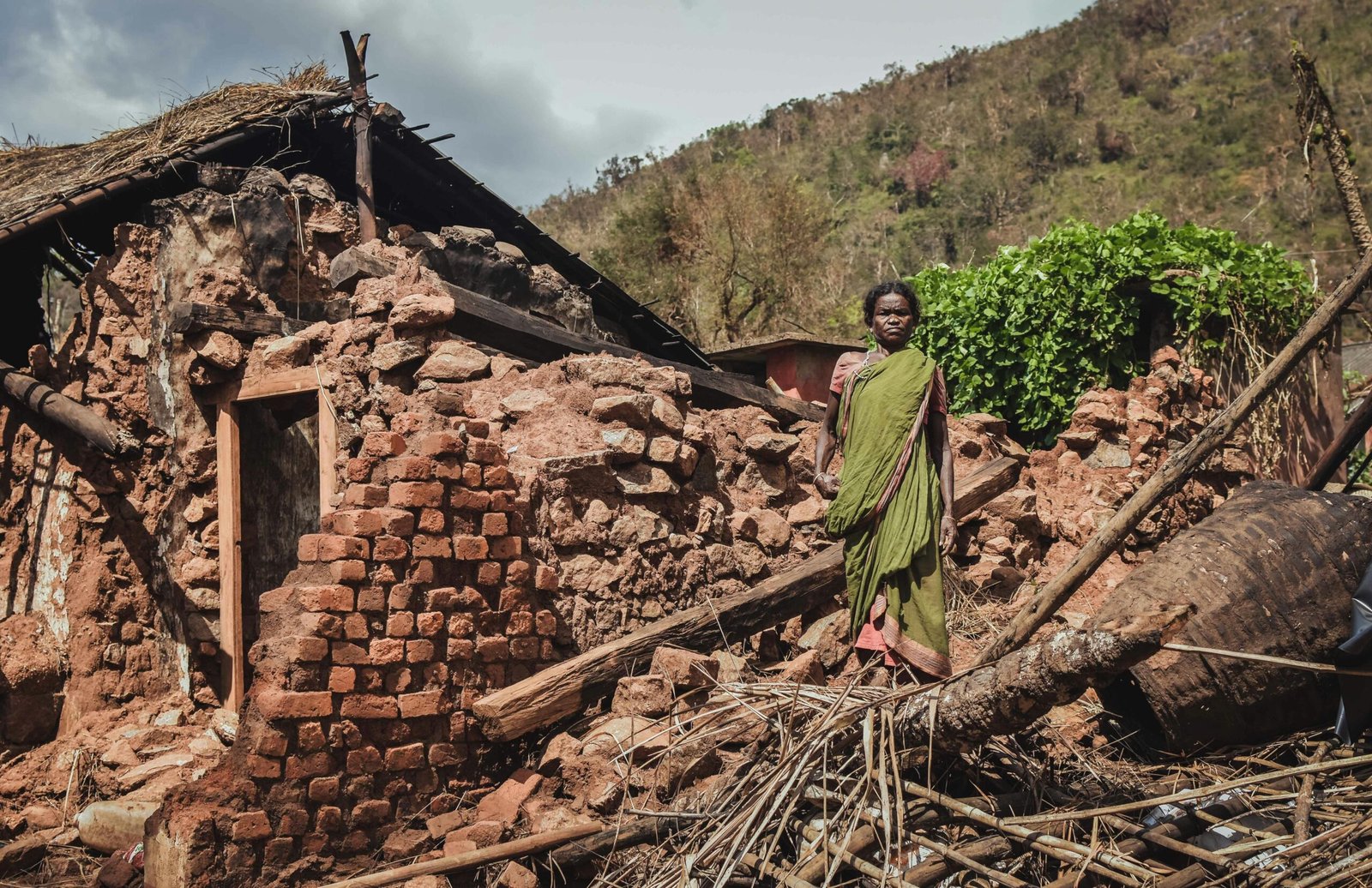 Woman stands in front of a destroyed house after a disaster in rural India, depicting struggle.