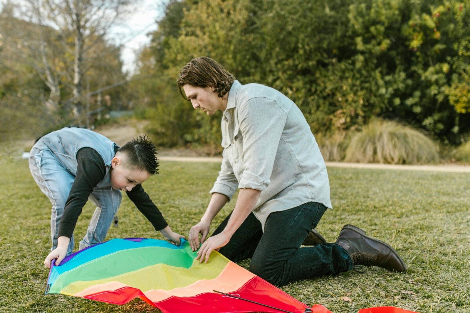 A father and son prepare a colorful kite on a grassy field, enjoying a sunny day outdoors.